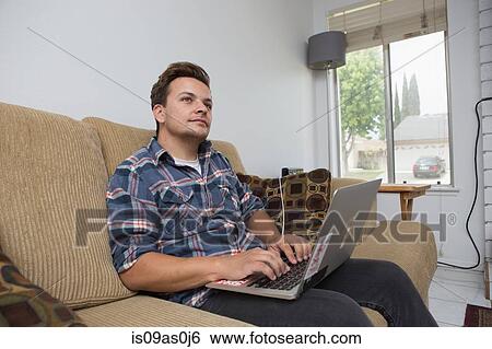 Young man sitting on sofa using laptop View Large Photo Image Stock Photograph - Young man sitting on sofa using laptop. Fotosearch