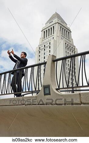 Stock Image - Business man standing on walkway using digital tablet to take photo, Los Angeles City Hall, California, USA. Fotosearch