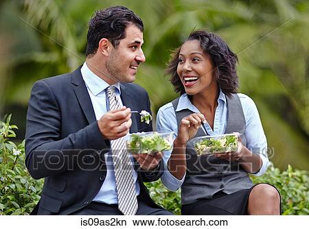 Picture - Business people sitting side by side enjoying a salad on lunch break, face to face, smiling. Fotosearch
