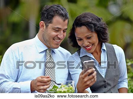 Picture - Business people sitting side by side enjoying a salad on lunch break, looking at smartphone, smiling. Fotosearch