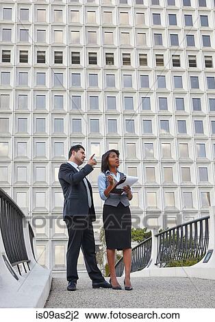 Stock Image - Business people standing talking holding paperwork, pointing and looking upwards. Fotosearch