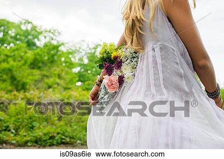 Stock Image - Cropped shot of young woman wearing white dress carrying bunch of flowers behind her back. Fotosearch