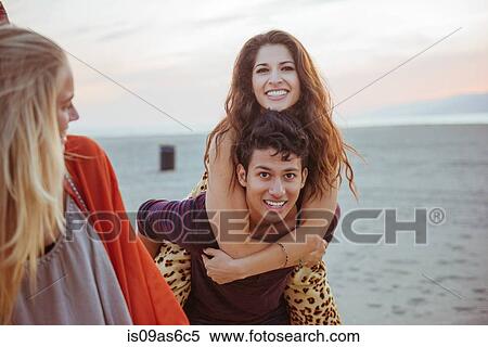 Stock Photography - Friends on beach, young man carrying young woman on back. Fotosearch