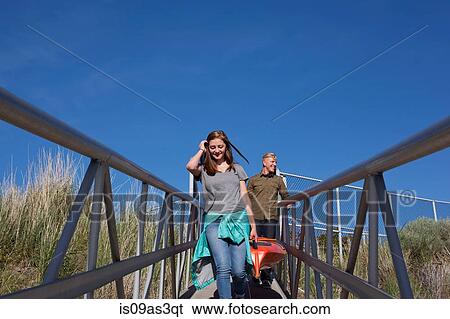 Stock Photograph - Front view of young couple carrying kayak on walkway with railings. Fotosearch