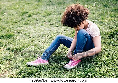Picture - Girl wearing pink shoes sitting on grass tying shoelace, looking down. Fotosearch