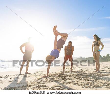 Stock Image - Group of friends on beach watching friend do handstand. Fotosearch