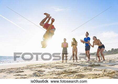 Stock Image - Group of friends on beach watching friend do somersault. Fotosearch