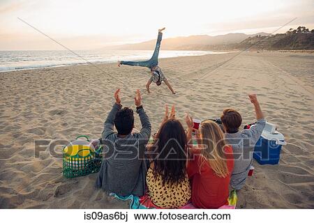 Group of friends sitting on beach, watching friend do cartwheels on and, sunset, rear view View Large Photo Image Stock Image - Group of friends sitting on beach, watching friend do cartwheels on and, sunset, rear view. Fotosearch