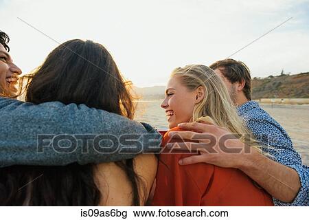 Stock Image - Group of friends sitting together on beach, at sunset, rear view. Fotosearch