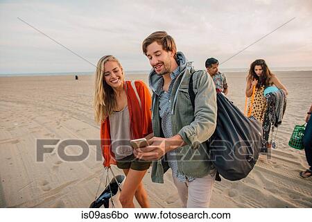 Picture - Group of friends walking along beach, young couple looking at smartphone. Fotosearch