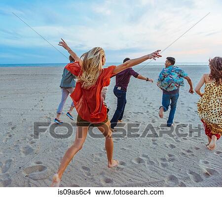 Picture - Group of friends walking along beach, young woman with arms in air, rear view. Fotosearch