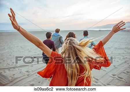 Stock Image - Group of friends walking along beach, young woman with arms in air, rear view. Fotosearch