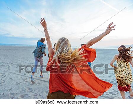 Stock Image - Group of friends walking along beach, young woman with arms in air, rear view. Fotosearch