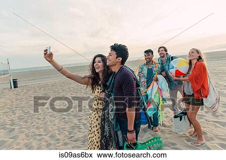 Stock Photo - Group of friends walking along beach, young couple taking self portrait with smartphone. Fotosearch