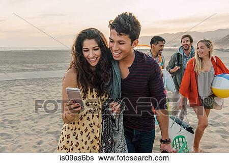 Group of friends walking along beach, young couple looking at smartphone View Large Photo Image Stock Photograph - Group of friends walking along beach, young couple looking at smartphone. Fotosearch