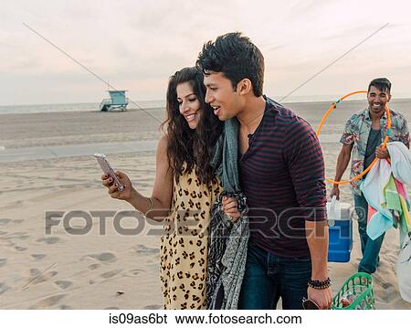 Group of friends walking along beach, young couple looking at smartphone View Large Photo Image Stock Photograph - Group of friends walking along beach, young couple looking at smartphone. Fotosearch