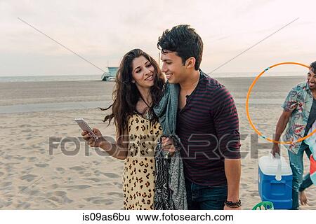 Stock Photograph - Group of friends walking along beach, young couple looking at smartphone. Fotosearch