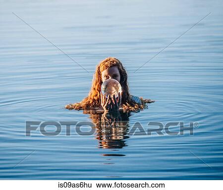 Stock Photograph - Head and shoulders of young woman with long red hair in lake holding crystal ball. Fotosearch