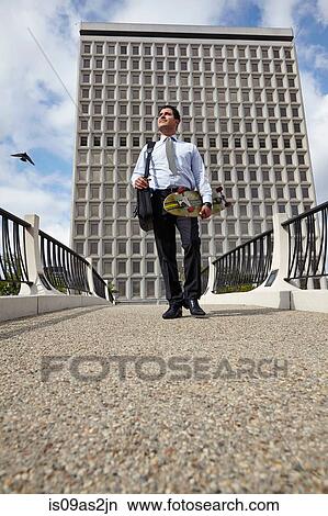 Picture - Low angle full length front view of mid adult business man carrying skateboard. Fotosearch