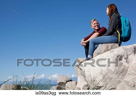 Stock Image - Low angle view of young couple sitting on rocks smiling. Fotosearch