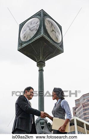 Low angle view side view of business people shaking hands underneath clock, waist up View Large Photo Image Picture - Low angle view side view of business people shaking hands underneath clock, waist up. Fotosearch