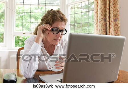 Stock Image - Mature woman drinking coffee whilst working on laptop in living room. Fotosearch