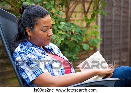 Stock Image - Mature woman on sun lounger reading book in garden. Fotosearch