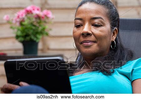 Stock Image - Mature woman sitting in garden reading digital tablet. Fotosearch