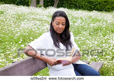 Stock Image - Mature woman sitting on park bench reading book. Fotosearch