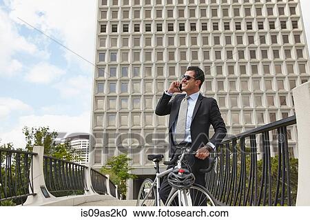 Stock Image - Mid adult business man holding bicycle, making telephone call using smartphone, wearing sunglasses. Fotosearch