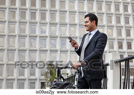 Mid adult business man holding bicycle, looking at smartphone, smiling View Large Photo Image Stock Photo - Mid adult business man holding bicycle, looking at smartphone, smiling. Fotosearch