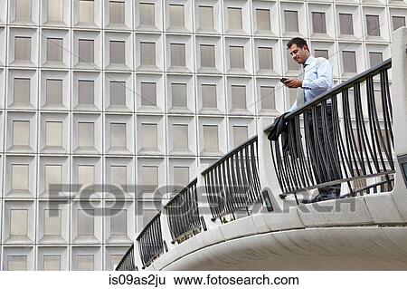 Stock Photograph - Mid adult business man standing on elevated walkway looking at smartphone. Fotosearch