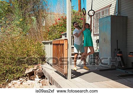 Stock Image - Mid adult man lifting daughter onto exercise rings on patio. Fotosearch