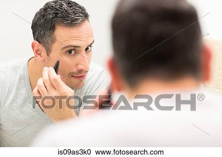 Stock Image - Mid adult man, looking in mirror, using tweezers, rear view. Fotosearch