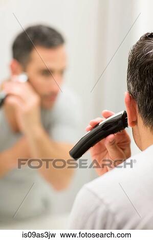 Stock Photo - Mid adult man, looking in mirror, using electric shaver, rear view. Fotosearch