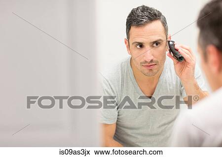 Stock Photo - Mid adult man, looking in mirror, using electric shaver. Fotosearch