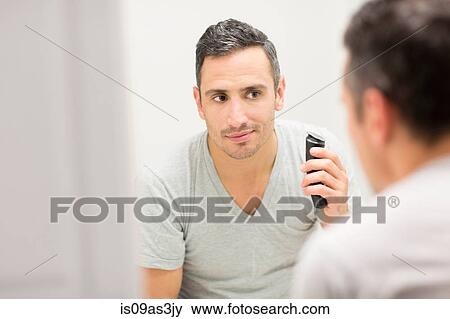 Stock Photo - Mid adult man, looking in mirror, using electric shaver. Fotosearch