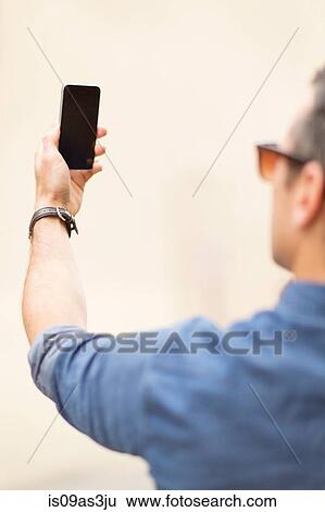 Stock Photograph - Mid adult man, outdoors, taking self portrait with smartphone, rear view. Fotosearch