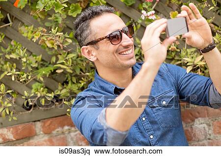 Stock Photography - Mid adult man, outdoors, taking self portrait with smartphone. Fotosearch