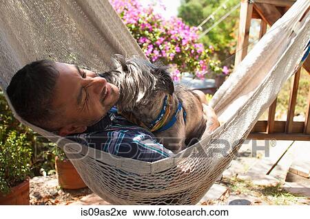 Stock Image - Mid adult man reclining in garden hammock with dog. Fotosearch