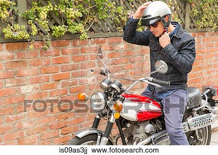 Stock Image - Mid adult man, sitting on motorbike, putting on helmet. Fotosearch