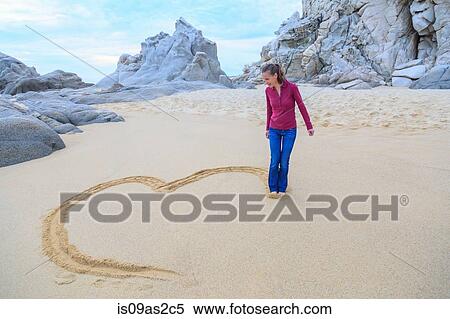 Stock Photography - Mid adult woman on beach, drawing heart shape with feet. Fotosearch