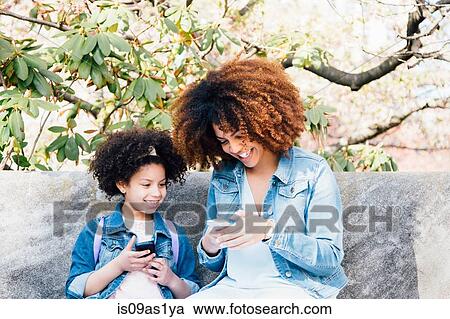 Mother and daughter sitting side by side looking at smartphones smiling View Large Photo Image Stock Image - Mother and daughter sitting side by side looking at smartphones smiling. Fotosearch