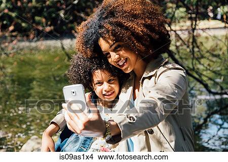 Picture - Mother with arm around daughter using smartphone to take selfie. Fotosearch