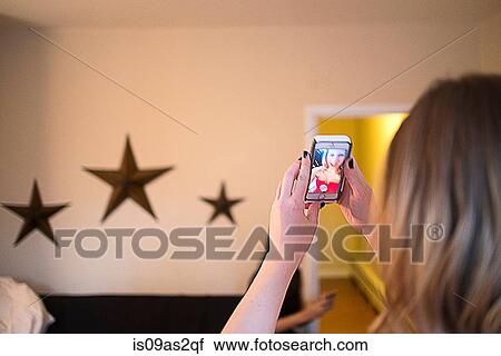 Stock Image - Over shoulder view of young woman taking smartphone selfie. Fotosearch