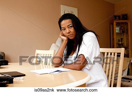 Stock Image - Portrait of fed up mature woman sitting at dining table reading bill. Fotosearch