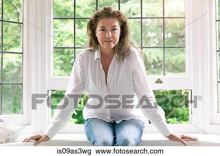 Stock Image - Portrait of mature woman sitting on living room windowsill. Fotosearch