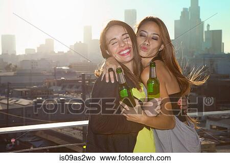 Picture - Portrait of two young women hugging at rooftop bar with Los Angeles skyline, USA. Fotosearch