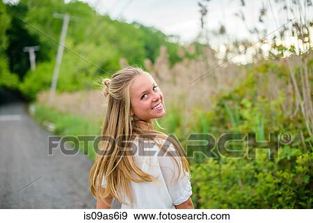 Stock Photo - Portrait of young woman with long blond hair looking back on rural road. Fotosearch