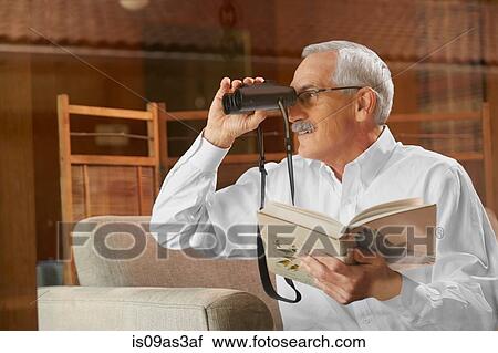 Stock Image - Senior man at home, birdwatching through window, using binoculars. Fotosearch